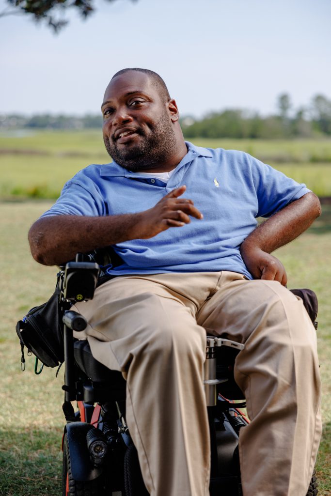 Photo of Alex, a Black man in a power wheelchair, speaking while outside with an SC lowcountry marsh behind him.