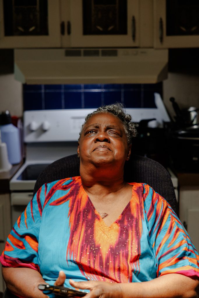 Photo of Susan, a black woman with a visual disability and curly cropped gray hair inside her home seated, smiling with a closed mouth.