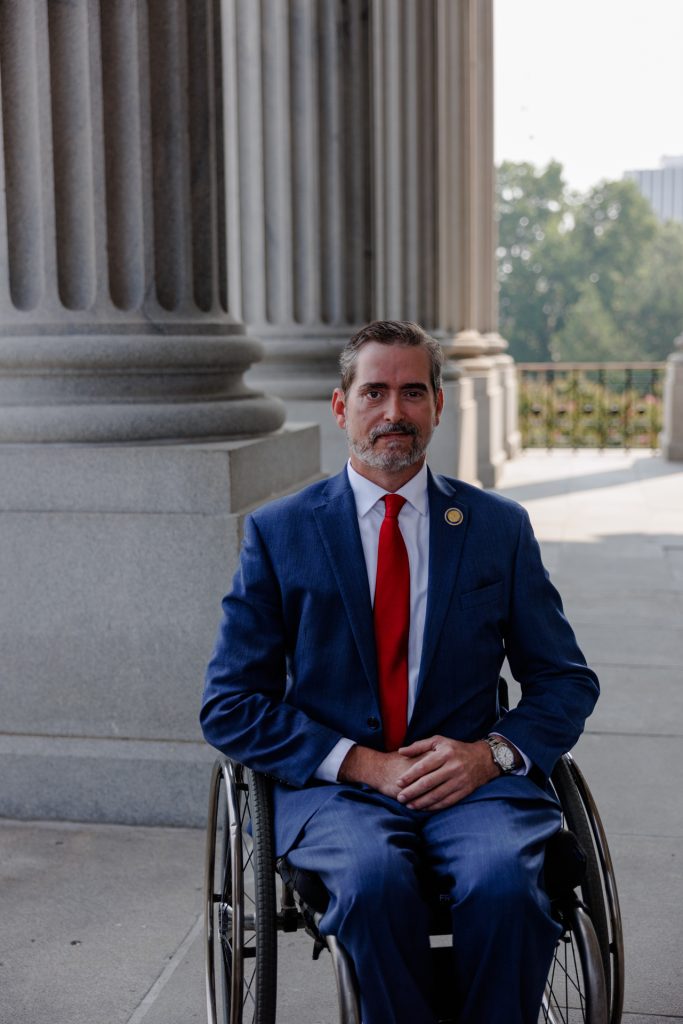 Photo of Cal, a white man in a suit with a red tie and SC house of representatives lapel pin, seated in his wheelchair while outside the SC statehouse under the large stone columns.