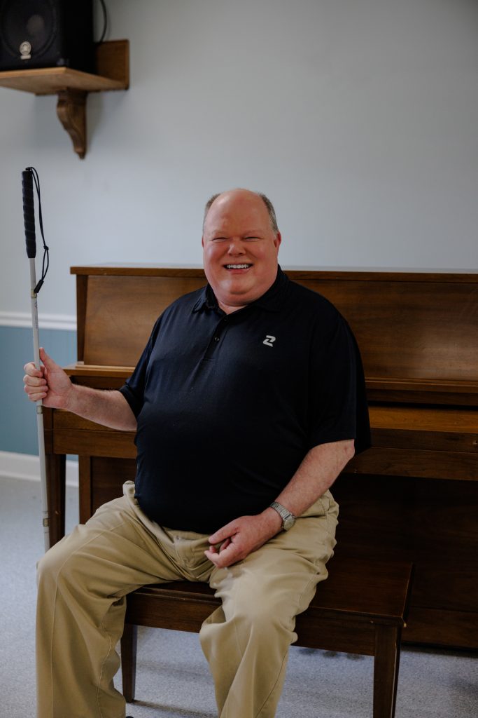 Photo of Marty, a white man who is blind, holding his long white cane and seated with an upright piano behind him.