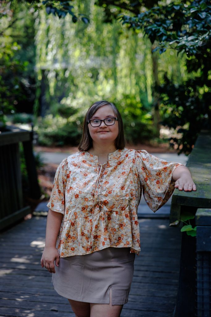 Photo of Brayden, a young white woman with down syndrome and glasses wearing a floral top and khaki skirt, posing on a wooden bridge in a green park shaded by trees.